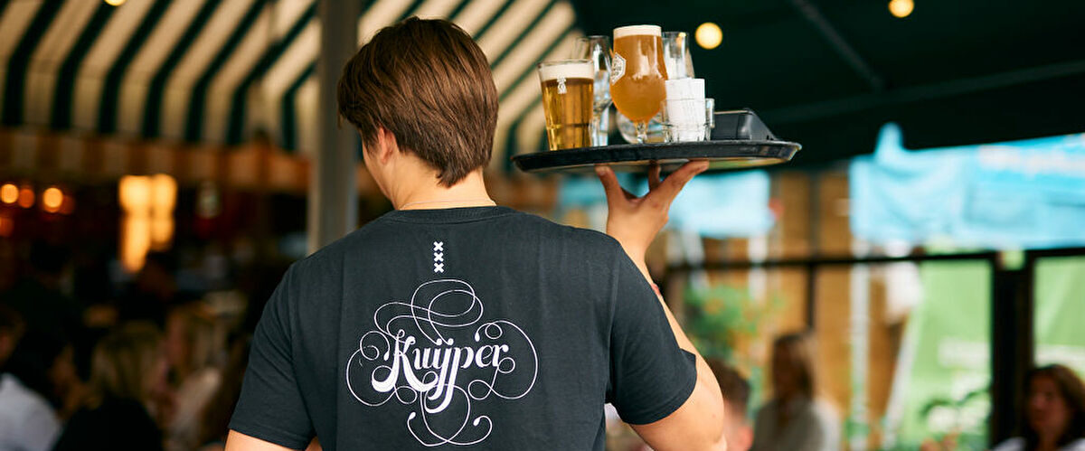 Bediening met een dienblad vol bier, in een Kuijper-shirt, onder de gestreepte luifel